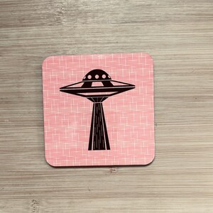 A single square pink coaster with a black flying saucer design featuring a wide base, on a light wood surface.