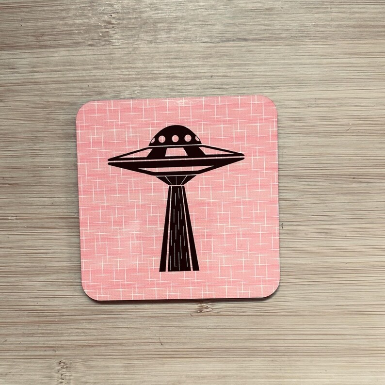 A single square pink coaster with a black flying saucer design featuring a wide base, on a light wood surface.