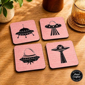 Four square pink coasters with black flying saucer designs, on a light wood surface next to a glass with a drink.