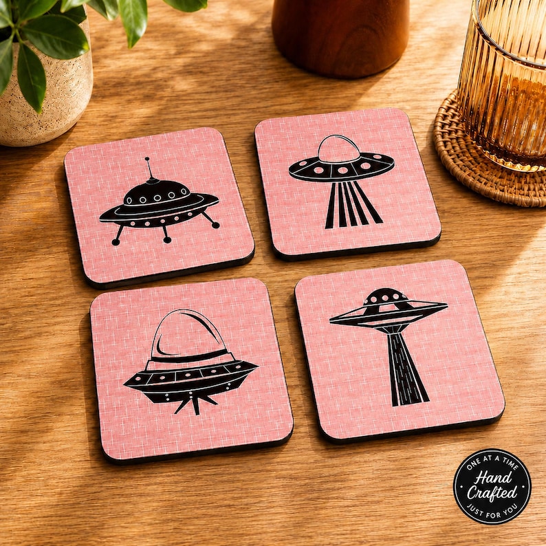 Four square pink coasters with black flying saucer designs, on a light wood surface next to a glass with a drink.