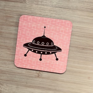 A single square pink coaster with a black flying saucer design featuring an antenna, on a light wood surface.