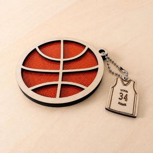 A circular wooden basketball ornament with a textured orange insert lies on a light wooden surface, attached by a chain to a jersey-shaped wooden tag.