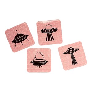 Four square pink coasters with black flying saucer designs, arranged in a diamond shape on a white background.
