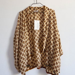 Puede incluir: Un kimono beige con estampado de lunares marrones y blancos. El kimono tiene cuello en V y mangas largas. Está colgado de una percha negra contra una pared blanca.