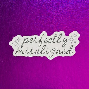 Op de afbeelding: Een witte sticker met de tekst "perfectly misaligned" in een handgeschreven lettertype. De sticker heeft een zwarte rand en is versierd met twee sterretjes. De achtergrond is een glanzend, paars oppervlak.
