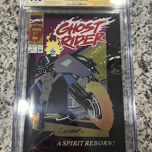 Op de afbeelding: Een gegradeerd stripboek in een beschermende hoes. De stripboekomslag toont een zwart-witte illustratie van Ghost Rider die op een motor rijdt. De titel van het stripboek is "Ghost Rider" en de tekst "A Spirit Reborn!" staat onderaan de omslag. Het stripboek is gegradeerd met 9.8 door CGC.