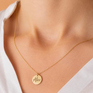 May include: A gold-toned necklace featuring a small, circular pendant with the name Alex engraved in a cursive script. The pendant is suspended from a delicate, thin gold-toned chain that rests against the collarbone area of the neck.