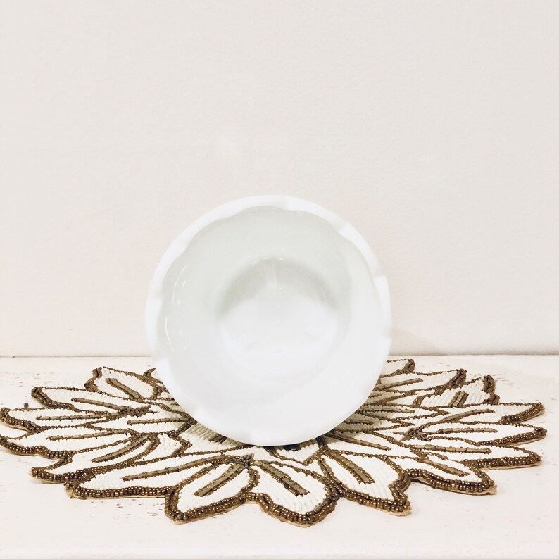 May include: White ceramic bowl with a scalloped edge, sitting on a beaded doily with a floral design.