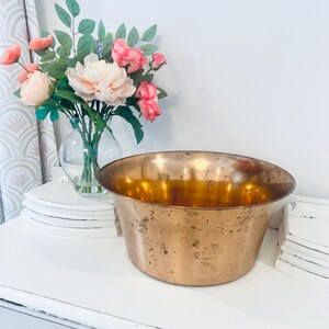 Puede incluir: Un cuenco grande de cobre antiguo con un borde ancho y un interior dorado y cálido. El cuenco está sobre una superficie blanca, con un ramo de flores rosas y melocotón en un jarrón de cristal en el fondo. El cuenco tiene un aspecto rústico y envejecido.