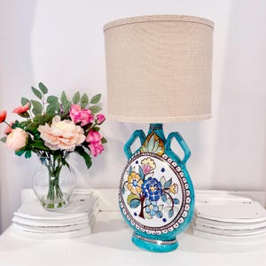 May include: A table lamp with a beige cylindrical fabric shade and a turquoise ceramic base shaped like a canteen with two handles. The base features a hand-painted floral design in blue, yellow, and green on a white circular center panel.