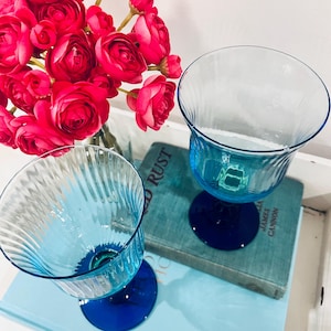 May include: Two clear, blue-tinted glass goblets with a ribbed design on the bowl and a dark blue base. A bouquet of bright pink flowers sits in the background. The goblets are on a light blue surface.