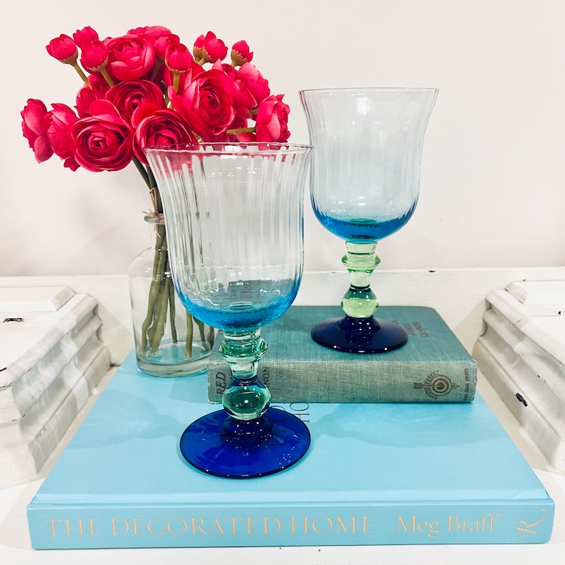 May include: Two clear glass goblets with blue and green accents. The goblets have a ribbed design and sit on a blue book. A vase of red flowers is in the background. The goblets have a blue base and rim, with a green stem.