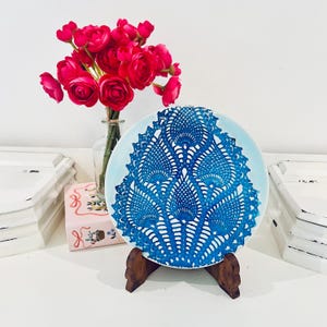 May include: A round white ceramic plate featuring a vibrant blue intricate lace-like pattern, displayed on a dark brown wooden stand. Beside the plate is a clear glass vase filled with a bouquet of bright pink artificial flowers.
