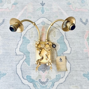 May include: Gold-toned wall sconce with a decorative bow design. The sconce features two curved arms holding light fixtures. A tag with the text "LIDIE ALIOTTI" is attached. The sconce is mounted on a patterned surface.