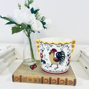 May include: A white ceramic planter with a rooster design, red, yellow, and black accents, and scalloped edges. The planter sits on a vintage book with a glass vase of white flowers in the background.