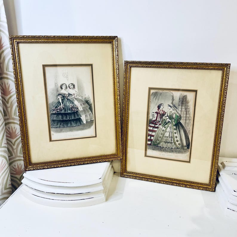 K&ouml;nnte beinhalten: Zwei gerahmte antike Drucke von Frauen in aufwendigen Kleidern. Die Drucke sind in Schwarzwei&szlig; mit Farbakzenten. Die Rahmen sind golden und verziert.