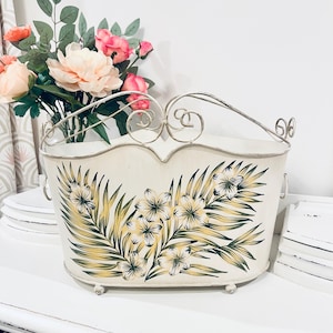 Könnte beinhalten: Cremefarbener Metallkorb mit einem floralen Design in Gelb und Grün. Der Korb hat einen geschwungenen Griff und kleine Füße. Ein Strauß rosa und pfirsichfarbener Blumen befindet sich hinter dem Korb.