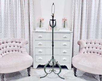 May include: A vintage floor lamp with a green metal base and a white lampshade. The lamp is standing in front of a white dresser with four drawers and two pink tufted armchairs.