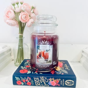 Op de afbeelding: Een grote glazen potkaars met een doorzichtig deksel, gevuld met dieprode was. Het etiket luidt "Yankee Candle Pomegranate Gin Fizz". De kaars staat op een blauw boek met bloemmotieven en de titel "Sense and Sensibility."