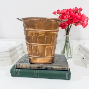May include: A metallic copper-colored bucket with a tapered shape and two small side handles, resting on top of two stacked hardcover books, one grey and one dark green, with a clear glass vase containing bright pink flowers nearby.