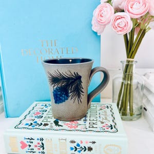 Puede incluir: Una taza de cerámica blanca con un asa negra y el texto "But First, Coffee" en negro, con una fuente de estilo manuscrito. La taza está sobre una superficie blanca.