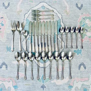 May include: A collection of stainless steel silverware, including forks, knives, and spoons, arranged on a patterned rug. The silverware has a decorative handle design. The set includes various sizes of spoons and forks, suitable for a formal dining setting.