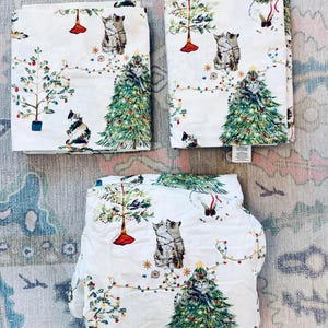 May include: Three folded white fabric pieces featuring a repeating holiday pattern of green Christmas trees, colorful string lights, and various cats. Some cats are sitting near trees, while others are depicted climbing or playing in the branches.