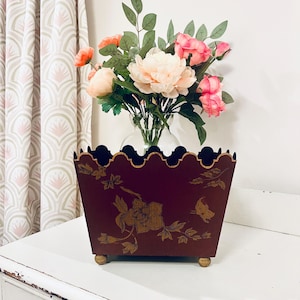以下が含まれることがあります： 金色の花と蝶のデザインが施された、バーガンディ色の長方形のプランター。 プランターは、金色のトリムが付いたスカラップのトップエッジがあり、4つの小さな丸い金色の足の上に置かれています。 ピンクとピーチの花で満たされたガラスの花瓶が中に入っています。