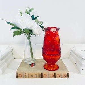 May include: A vibrant red glass owl-shaped vase with a textured surface. The vase sits on a vintage book next to a clear glass vase holding white flowers. The vase has a sticker that says "Viking".