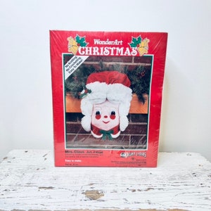 Peut inclure: Un kit de loisirs créatifs WonderArt de Noël vintage représentant une tête de Mme Claus. Le kit est rouge et blanc avec une image de l'artisanat fini sur le devant. Le texte sur le devant indique "WonderArt Christmas", "No Knitting. No Crocheting. Easy To Make!" et "Mrs. Claus Art. #K136 Easy to make. Net wt. 15 oz."