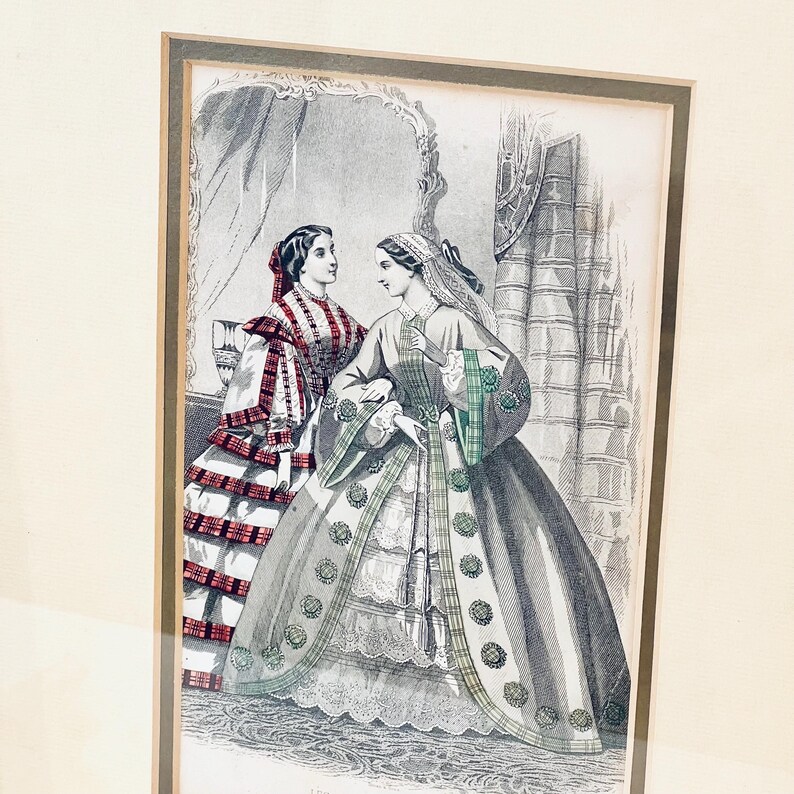 K&ouml;nnte beinhalten: Ein Vintage-Schwarzwei&szlig;druck von zwei Frauen in aufwendigen Kleidern. Die Frau links tr&auml;gt ein rot-wei&szlig; kariertes Kleid mit einer schwarzen Sch&auml;rpe. Die Frau rechts tr&auml;gt ein graues Kleid mit gr&uuml;n-schwarzen Karomustern. Beide Frauen stehen in einem Raum mit einem Spiegel und einem Fenster.