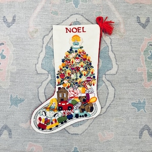Puede incluir: Un calcetín navideño blanco con diseños bordados de un árbol decorado con un ángel en la punta, una casa, un oso de peluche, un tren de juguete y regalos. La palabra NOEL está bordada en rojo en la parte superior y tiene una borla roja.