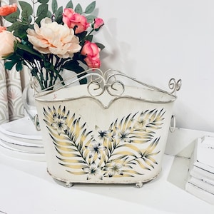 Könnte beinhalten: Cremefarbener Metallpflanzer mit einem floralen Design in Gelb und Grün. Der Pflanzer hat einen dekorativen, gerollten Metallgriff und Füße. Ein Strauß rosa und pfirsichfarbener Blumen befindet sich im Inneren.