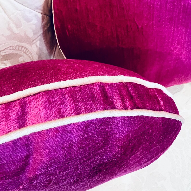 May include: Close-up of two plush, magenta velvet pillows with white piping. The pillows are in a decorative setting, with a soft, textured background. The pillows have a luxurious, inviting appearance.