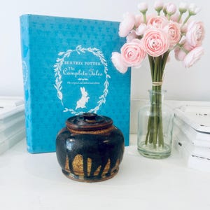 Puede incluir: Una taza de cerámica blanca con asa negra y el texto "But First, Coffee" en negrita negra. La taza se encuentra sobre un fondo borroso, sugiriendo un ambiente acogedor.