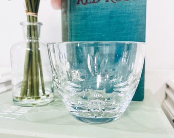 Puede incluir: Un cuenco de cristal tallado transparente con un patrón floral decorativo. El cuenco está delante de un libro turquesa titulado "RED RUST" y un jarrón de cristal con flores rosas. Los artículos están sobre un libro verde claro.