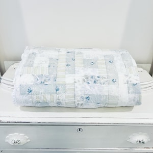 May include: A folded patchwork quilt with a mix of white, light blue, and gray squares. Some squares feature floral patterns. The quilt has a white lace trim and is placed on a white surface. A cozy home decor item.