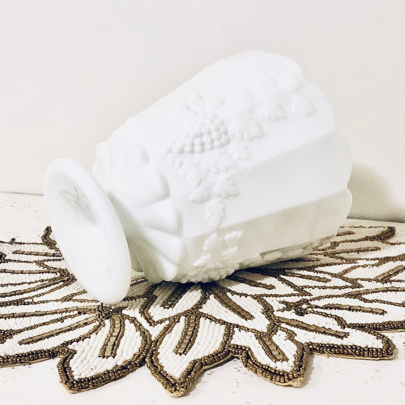 May include: White milk glass vase with a grapevine design. The vase is sitting on a gold and white beaded doily.