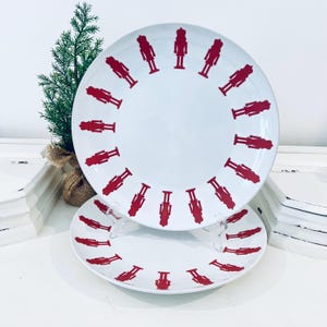 Puede incluir: Dos platos redondos de cerámica blanca con un diseño de cascanueces rojo. Los platos presentan un anillo de figuras de cascanueces rojas alrededor del borde. Un pequeño árbol verde en una maceta de arpillera está en el fondo.