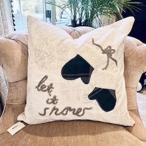 May include: White decorative pillow with a snowflake pattern and the words "let it snow" embroidered in brown. Features two green plaid mittens and a decorative bow. The pillow is on a beige upholstered chair.