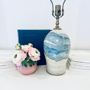 Puede incluir: Una lámpara de mesa de cerámica con un diseño veteado azul y blanco. La lámpara tiene una base de latón y un cuerpo de cerámica color crema. Un pequeño jarrón rosa con flores rosas está al lado de la lámpara.