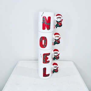 Puede incluir: Cuatro tazas de cerámica blanca apiladas una encima de la otra. Cada taza tiene una letra roja en la parte delantera, deletreando la palabra "NOEL". Cada taza tiene una pequeña figura de elfo unida al asa.