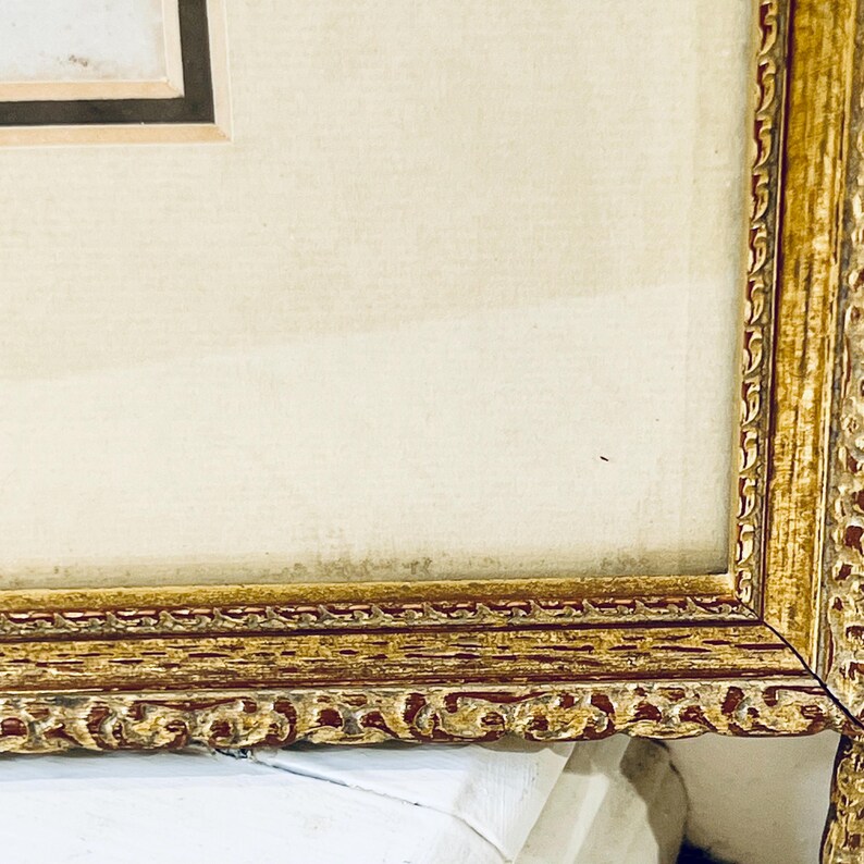 K&ouml;nnte beinhalten: Ein goldfarbener Bilderrahmen mit einem detaillierten, verzierten Design. Der Rahmen ist aus Holz und hat einen hellbraunen Passepartout.