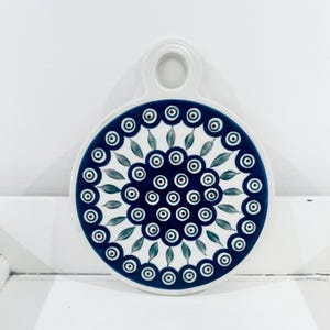 May include: A round ceramic serving board with a handle. The board has a dark blue border with a repeating floral pattern in white, green, and red. The handle has a circular opening. The board is white.