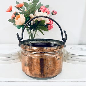 May include: A cylindrical copper pot with a weathered, tarnished finish and a dark metal swing handle attached to two side hooks. The pot features horizontal ridges along the upper rim and shows signs of oxidation and surface wear throughout.