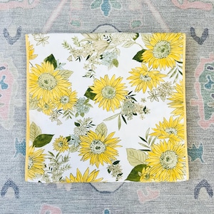 May include: A white tablecloth with a yellow border and a repeating pattern of yellow sunflowers and green leaves.