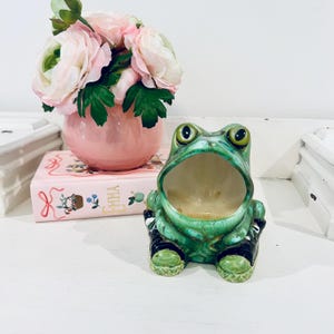 May include: A green ceramic frog figurine with a wide open mouth, positioned in front of a pink book titled Emma and a pink vase filled with light pink artificial flowers. The frog has dark green accents and textured skin details.