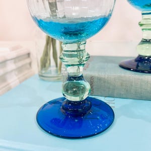 May include: Two elegant, hand-blown glass goblets with a vibrant blue and green color scheme. The bowls are filled with a turquoise liquid. The stems feature a unique, rounded design, and the bases are a deep cobalt blue. The glasses sit on a light blue surface.