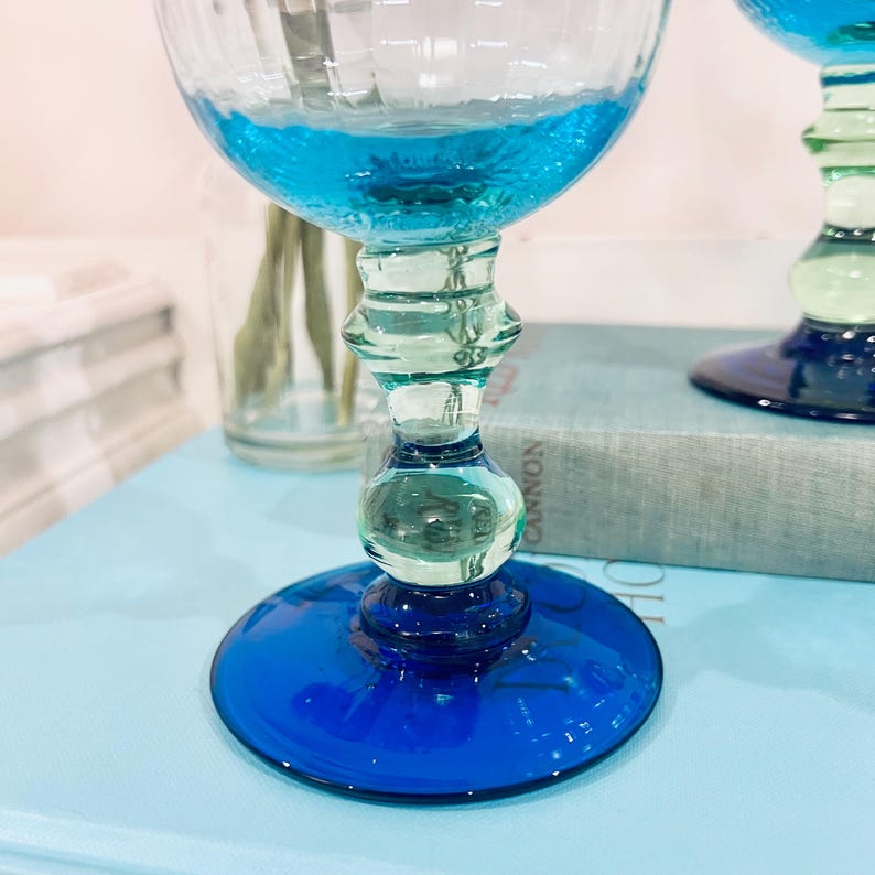 May include: Two elegant, hand-blown glass goblets with a vibrant blue and green color scheme. The bowls are filled with a turquoise liquid. The stems feature a unique, rounded design, and the bases are a deep cobalt blue. The glasses sit on a light blue surface.