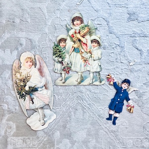 Könnte beinhalten: Vintage Weihnacht Engel-Ausschnitte. Ein Engel hält einen Weihnachtsbaum, während andere Trompeten und Glocken halten. Die Engel sind in weißen und blauen Outfits mit goldenen Akzenten und Flügeln gekleidet. Die Ausschnitte befinden sich auf einem strukturierten, hellgrauen Hintergrund.